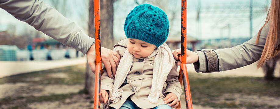 Child Custody - a baby is in a swing. A male hand is holding onto the right arm and a female hand is holding onto the left arm. The child is not smiling and looks sad.